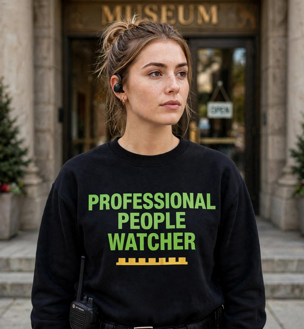 Woman modeling the black People Watcher sweatshirt at a museum entrance, green print PROFESSIONAL PEOPLE WATCHER with gold battlement strip fully visible
