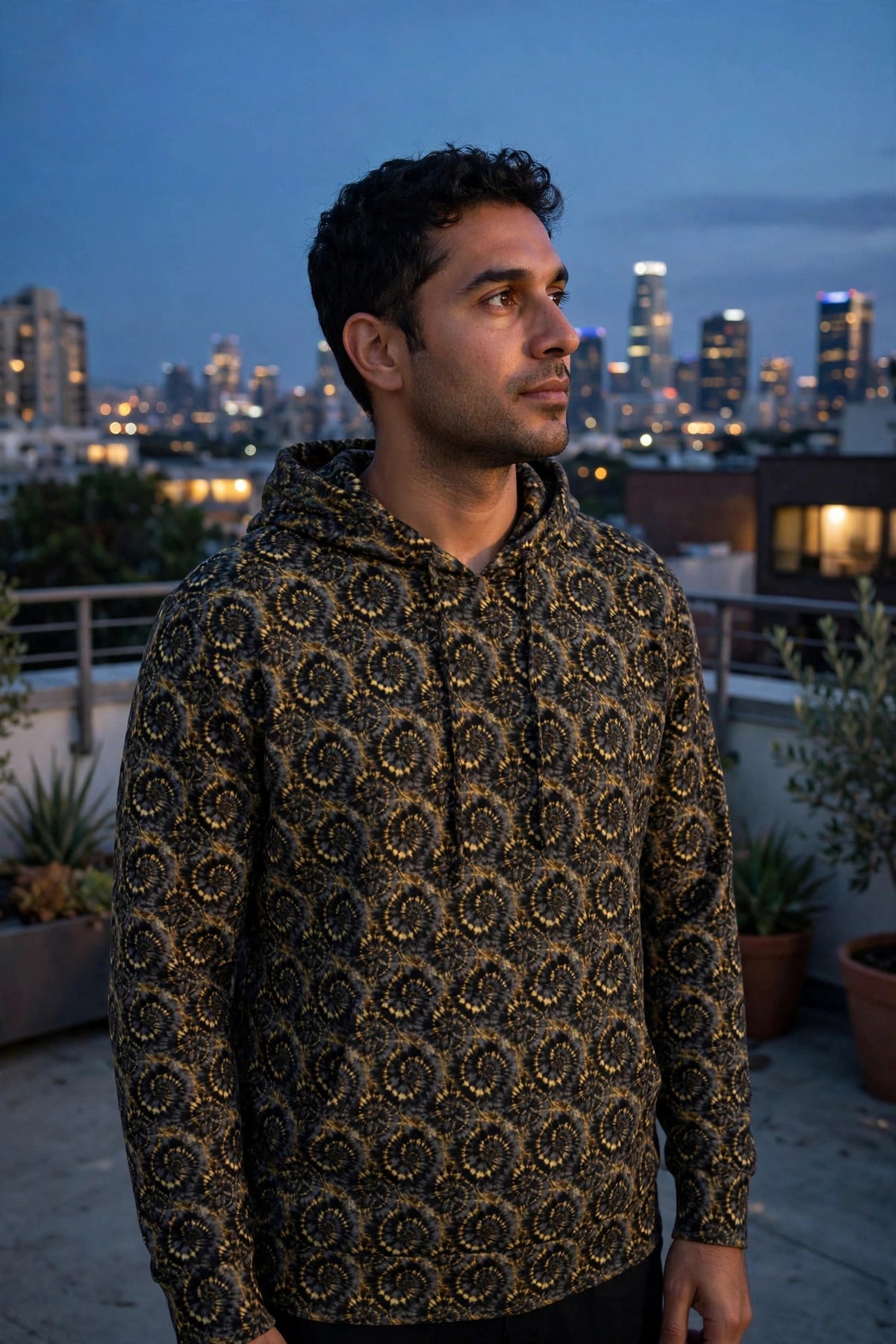 Man wearing the black and gold spiral pattern Rooftop hoodie on a terrace at dusk with a skyline behind him