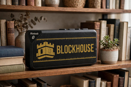 Blockhouse Bluetooth speaker with gold tower and BLOCKHOUSE print on a living room bookshelf beside books and a plant