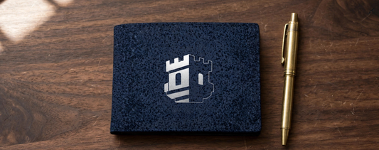 Citadel bifold wallet closed on a wooden desk beside a brass pen, showing the silver tower emblem on the dark speckled exterior.