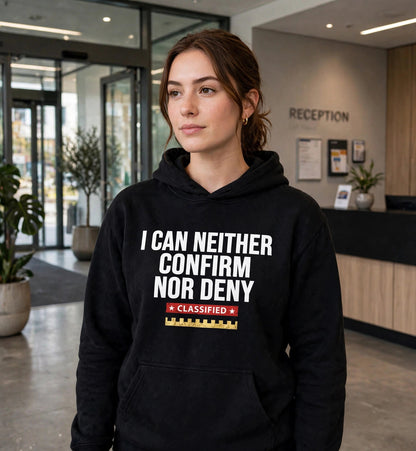 Woman modeling the black Confirm Nor Deny hoodie at an office reception, white print I CAN NEITHER CONFIRM NOR DENY with red CLASSIFIED strip and gold battlement fully visible