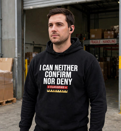 Man modeling the black Confirm Nor Deny hoodie at a warehouse loading dock, white print I CAN NEITHER CONFIRM NOR DENY with red CLASSIFIED strip and gold battlement fully visible