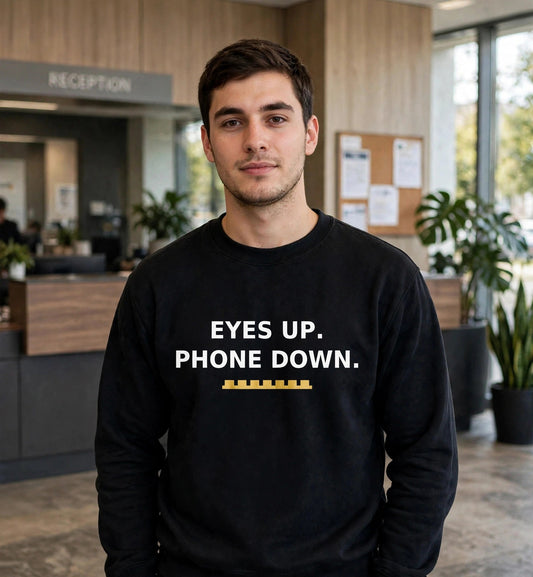 Man modeling the black Eyes Up sweatshirt in an office reception lobby, white print EYES UP. PHONE DOWN. with gold battlement strip fully visible