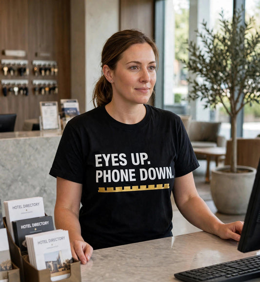 Woman modeling the black Eyes Up t-shirt at a hotel desk, white text EYES UP. PHONE DOWN. fully visible