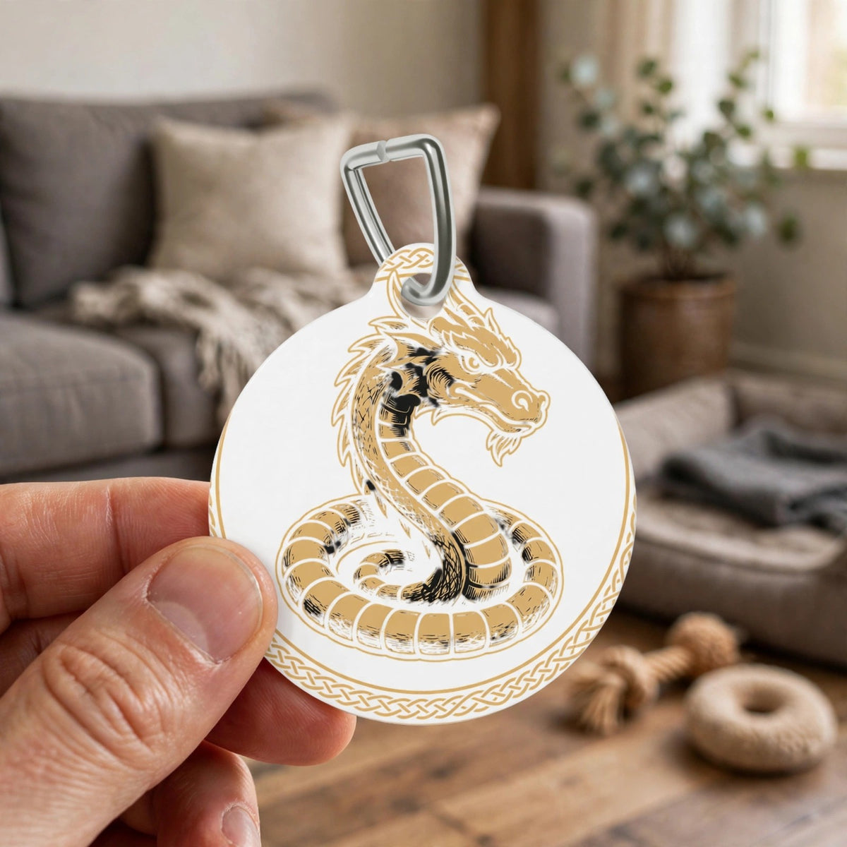 Hand holding the Fáfnir Head Mark pet ID tag in a living room, gold dragon design facing the camera