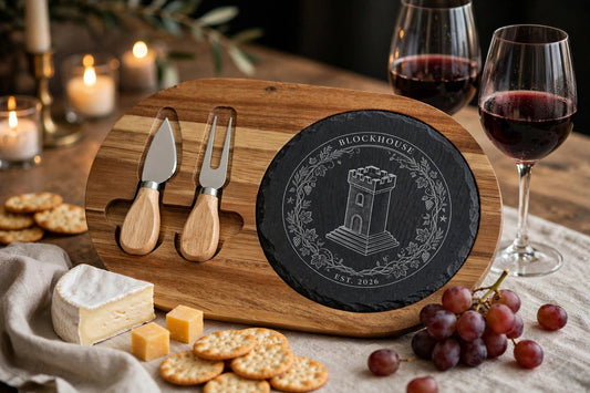 Feast Crest charcuterie board on a styled table — acacia wood board with dark charcoal slate insert engraved with the Blockhouse tower crest, cheese, grapes and red wine