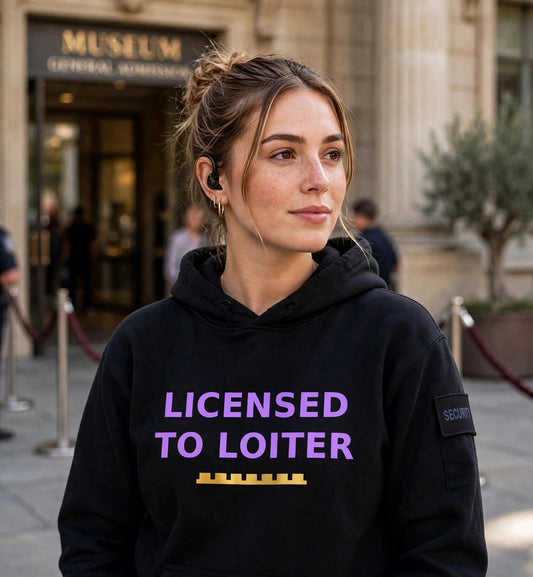 Woman modeling the black Licensed To Loiter hoodie at a museum entrance, purple print LICENSED TO LOITER with gold battlement strip fully visible