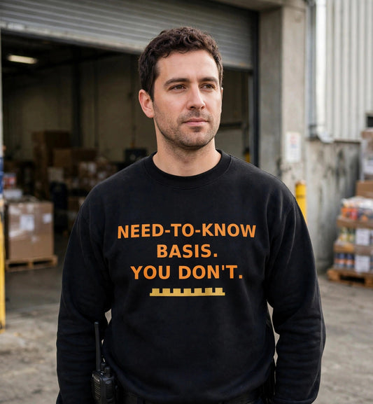 Man modeling the black Need To Know sweatshirt at a warehouse loading dock, orange print NEED-TO-KNOW BASIS. YOU DON'T. with gold battlement strip fully visible
