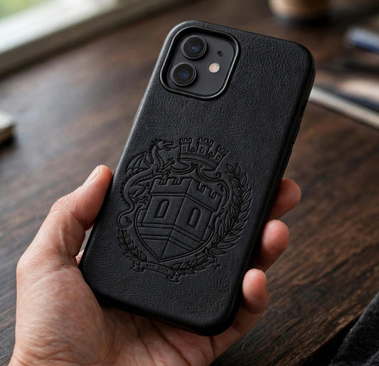 The Sovereign Crest black leather phone case held in a hand over a dark wood desk, showing the embossed crest on the case back