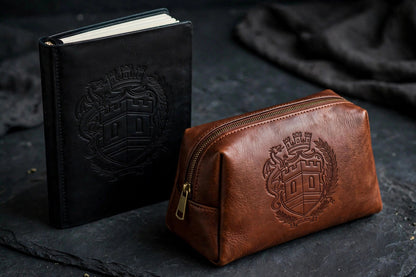 The Sovereign Crest Leather Set Two on dark slate seen from a closer angle: black leather journal and brown leather toiletry bag with the embossed crest