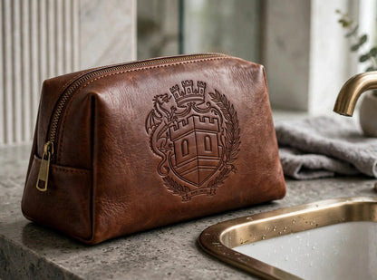 The Sovereign Crest brown leather toiletry bag on a bathroom counter beside a sink, showing the embossed crest