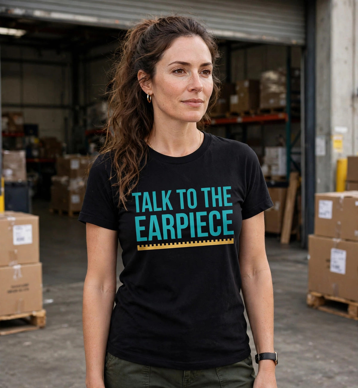 Woman wearing the black Talk To The Earpiece t-shirt in teal lettering in a warehouse loading area