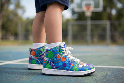 Side view of a child wearing the Tower Explosion kids' high-top basketball sneakers on an outdoor basketball court — electric-blue high-tops with a multicolor paint-splatter explosion print, white laces and white soles.