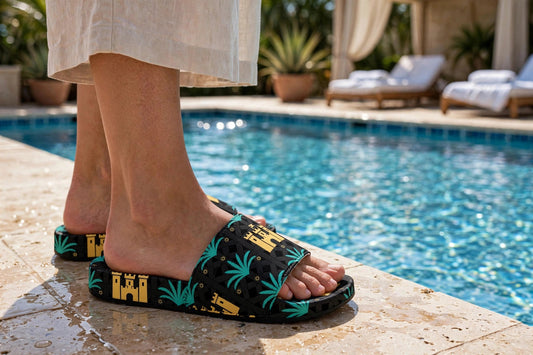 Woman wearing Blockhouse colorful tropical print slide sandals, side view, standing on a pool deck - black slides with teal palm fronds, gold castle and small gold suns